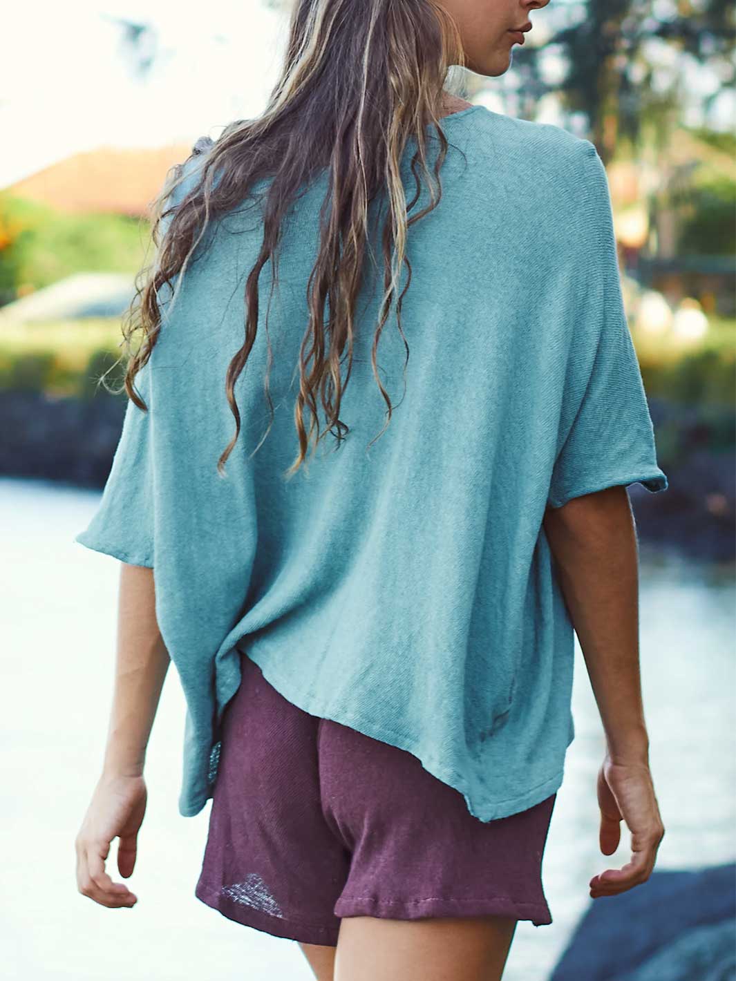 A woman shown from behind wearing a loose, short-sleeve teal top and mauve-purple shorts, standing outdoors near water with a blurred natural background.