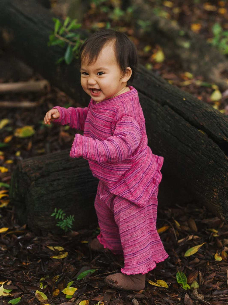 A smiling toddler standing outdoors on a forest floor, wearing a bright pink textured long-sleeve top and matching pants.