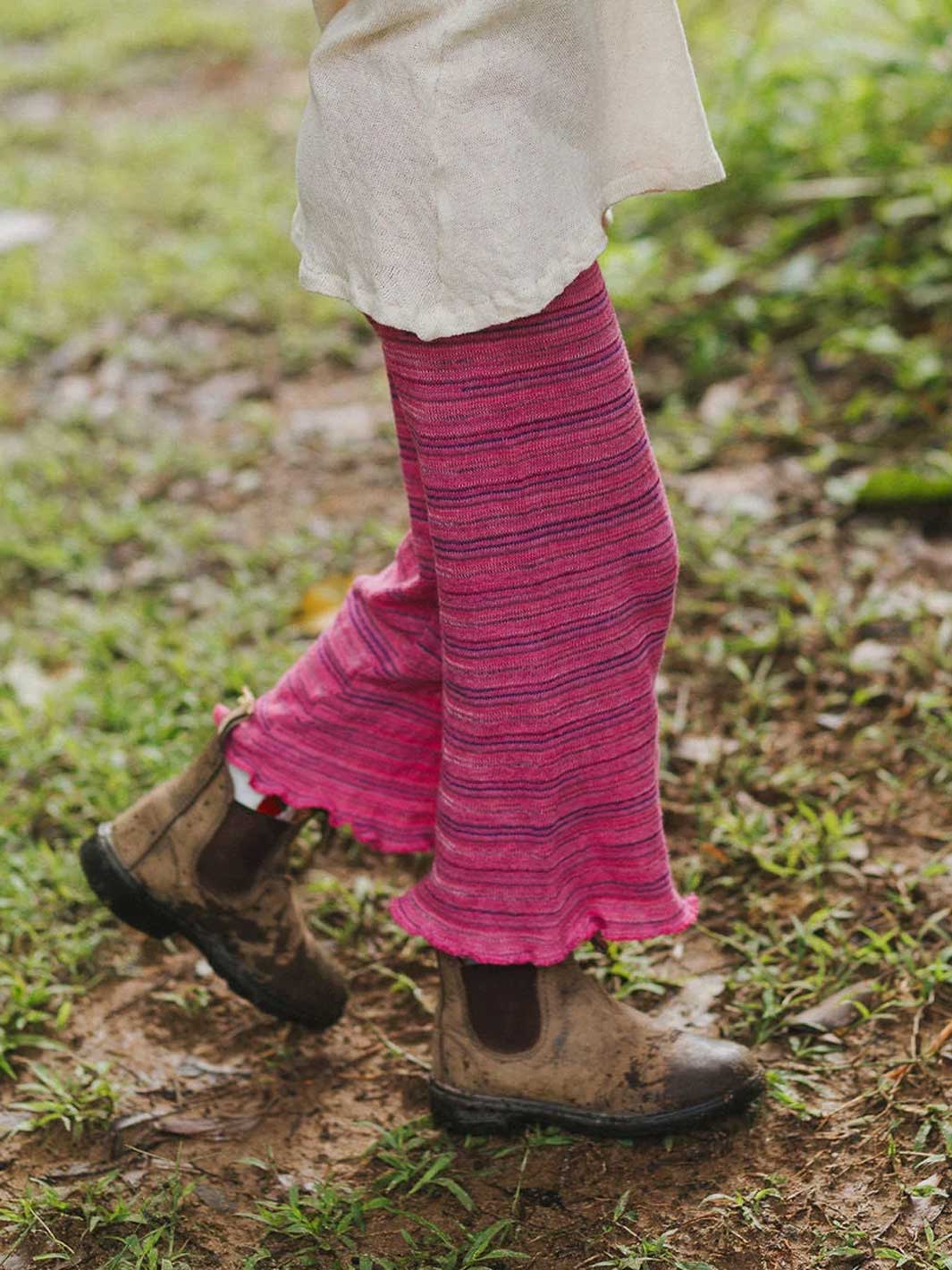 A person outdoors wearing bright pink striped knit cropped pants with ruffled hems, paired with brown boots on a muddy grassy ground.
