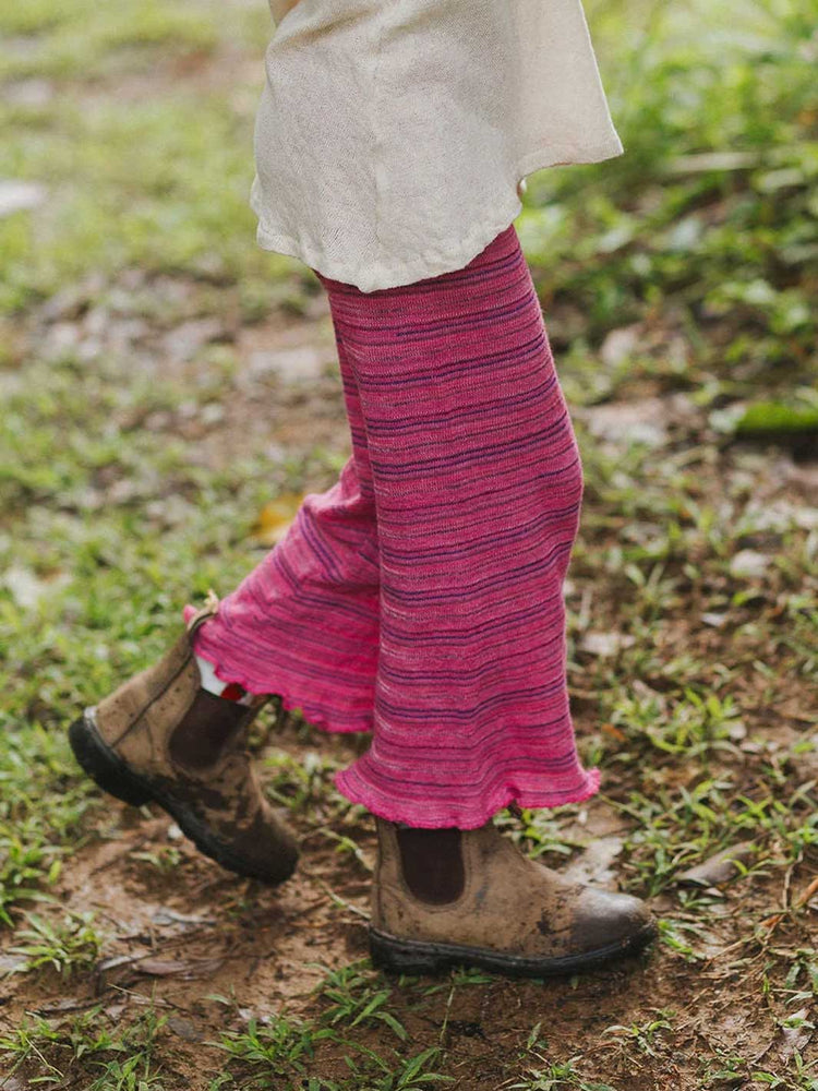 A person outdoors wearing bright pink striped knit cropped pants with ruffled hems, paired with brown boots on a muddy grassy ground.