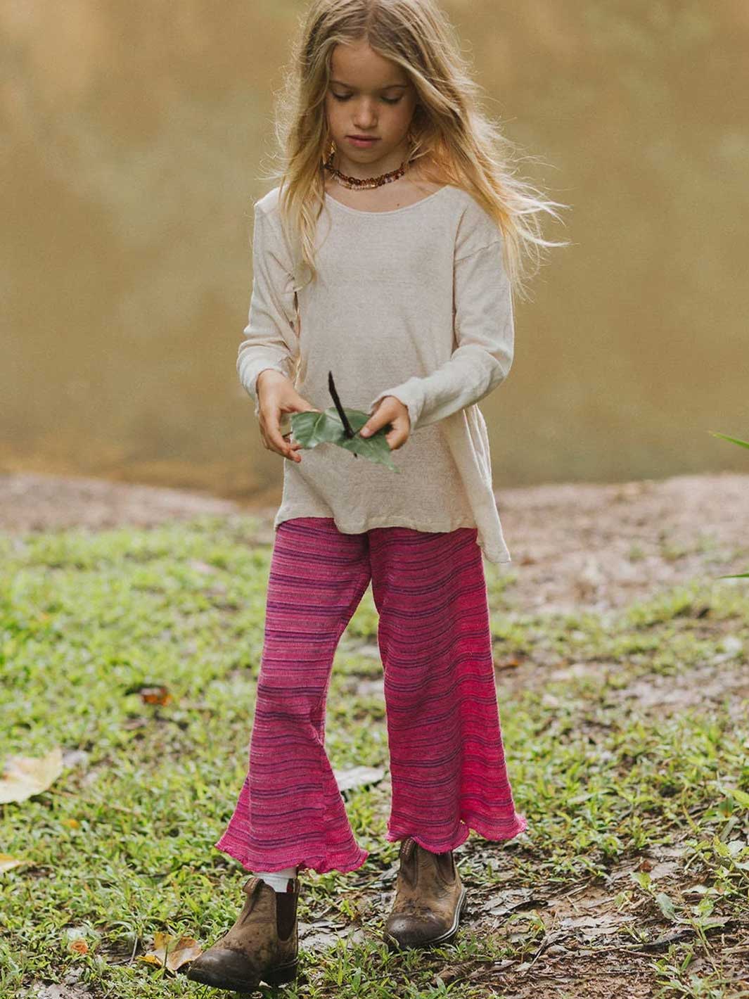 A young girl standing outdoors on grass and dirt, wearing a light beige long-sleeve top and bright pink striped flared pants with brown boots.