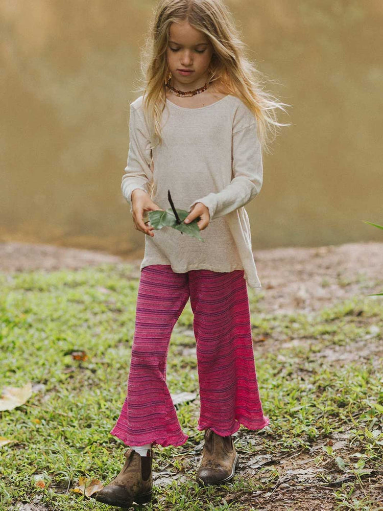 A young girl standing outdoors on grass and dirt, wearing a light beige long-sleeve top and bright pink striped flared pants with brown boots.