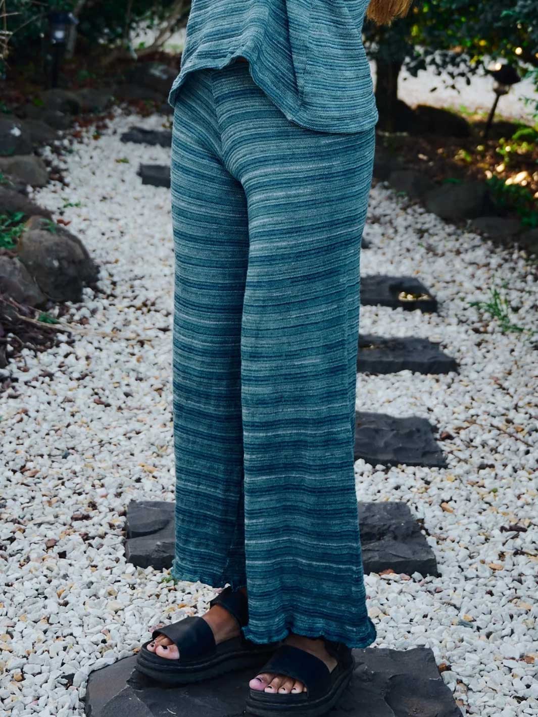 A person wearing teal and blue striped wide-leg pants and black slide sandals, standing outdoors on a stone path covered in white gravel.