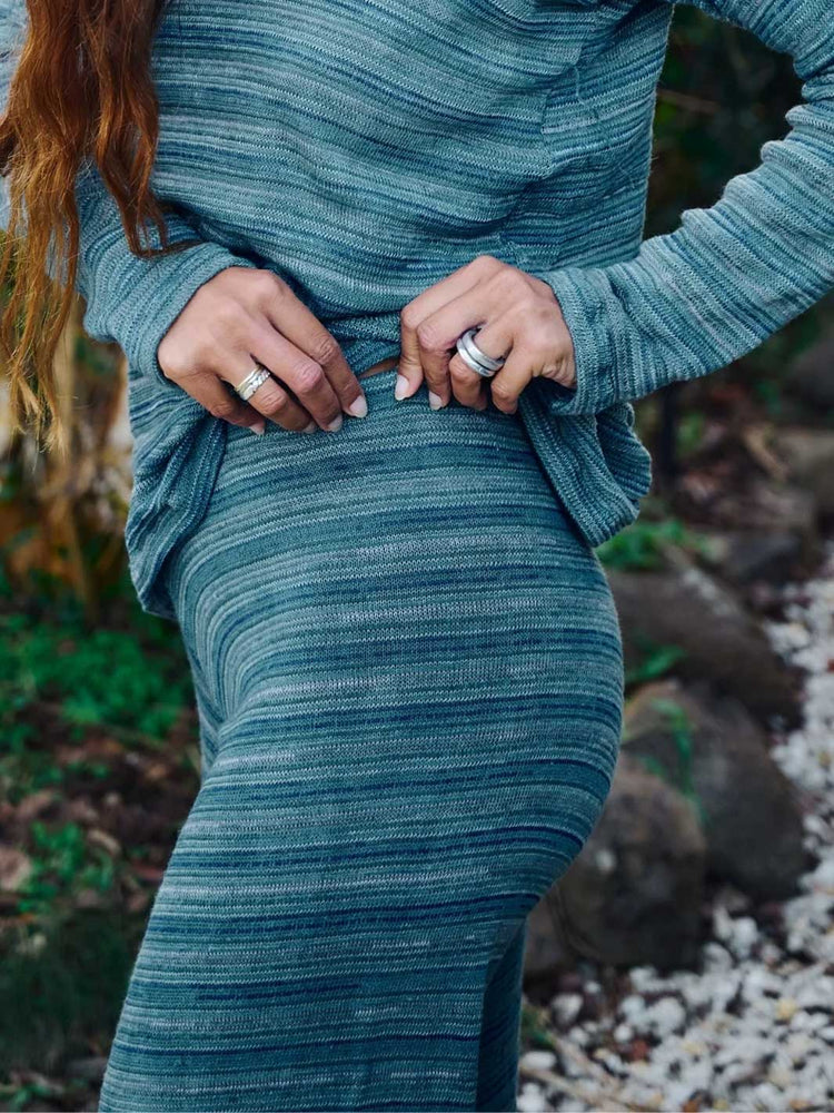 A close-up of a person wearing a ribbed, long-sleeve knit top and matching pants in a muted teal-blue colour, standing outdoors with a blurred natural background of rocks and foliage.