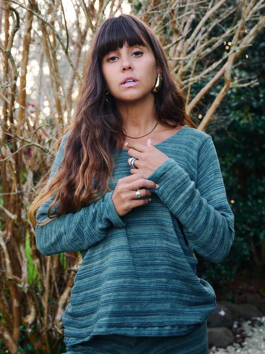 A woman with long brown hair and bangs wearing a textured, heathered teal-green long-sleeve top, standing outdoors in front of a background of dry branches and green foliage.