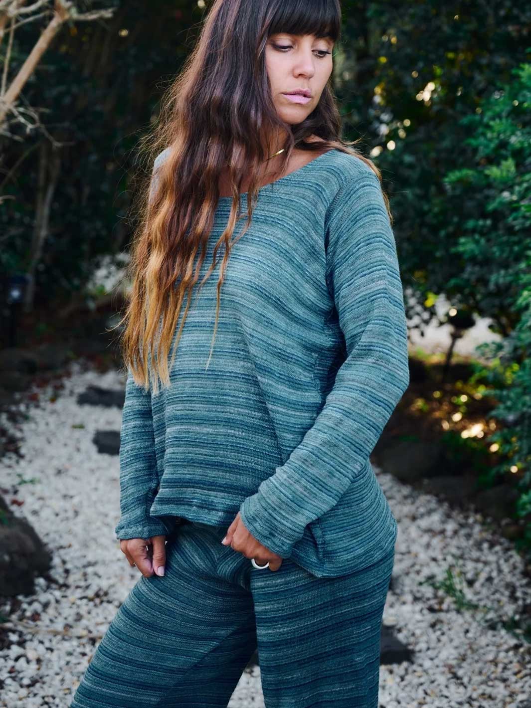 A woman wearing a matching two-piece loungewear set in a textured teal-green heathered fabric, standing outdoors on a gravel path with green foliage in the background.