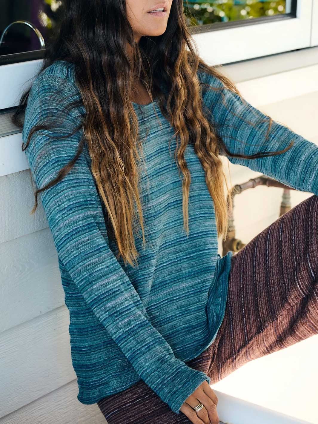 A woman wearing a long-sleeve textured knit top in a heathered teal blue color, paired with brown striped pants, sitting outdoors against a white wall.