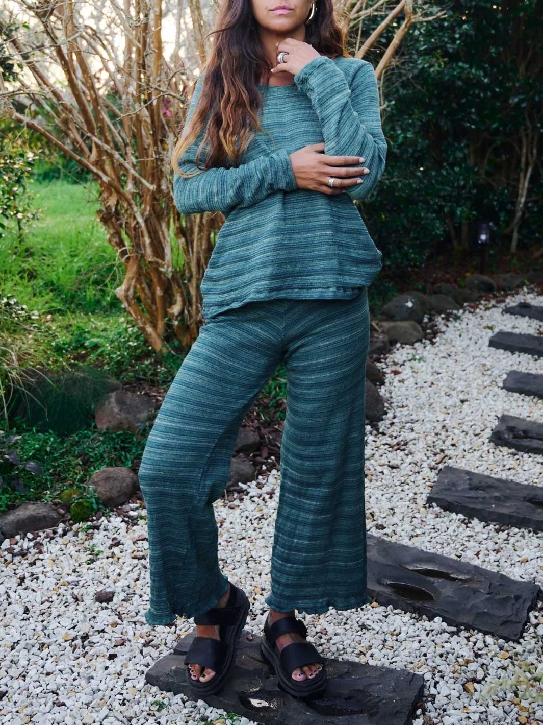 A woman wearing a matching two-piece ribbed knit lounge set in a muted teal green, standing outdoors on a stone path surrounded by white gravel and greenery.