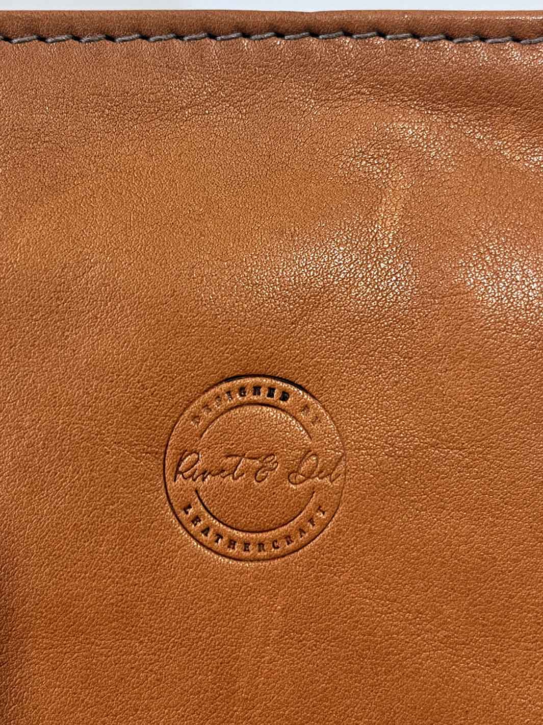 Close-up of a tan leather clutch bag or pouch showing the embossed circular 'Rivet & Del' logo and stitched edge at the top.