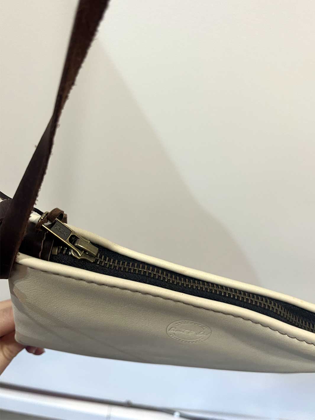 Close-up of a cream-colored leather crossbody bag with a dark brown strap and a gold-toned zipper, held by a hand against a plain light gray background.