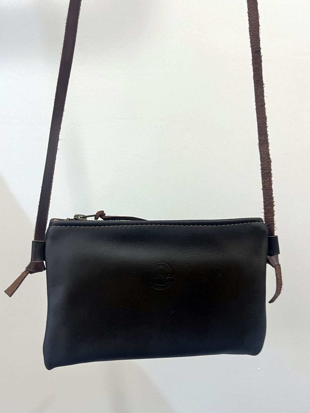 A black leather crossbody bag with a brown leather strap and zipper pull, featuring a subtle embossed logo on the front, shown against a plain white background.
