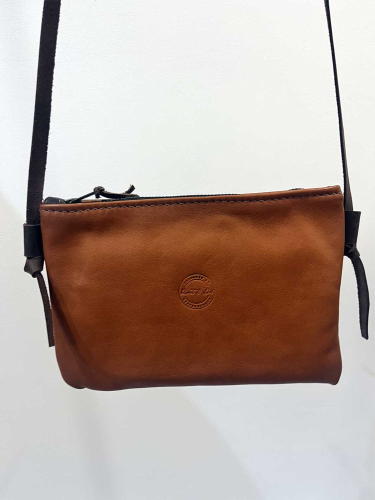 A tan brown leather crossbody bag with a dark brown adjustable strap, featuring a circular embossed logo on the front and a zippered top closure, shown against a plain white background.