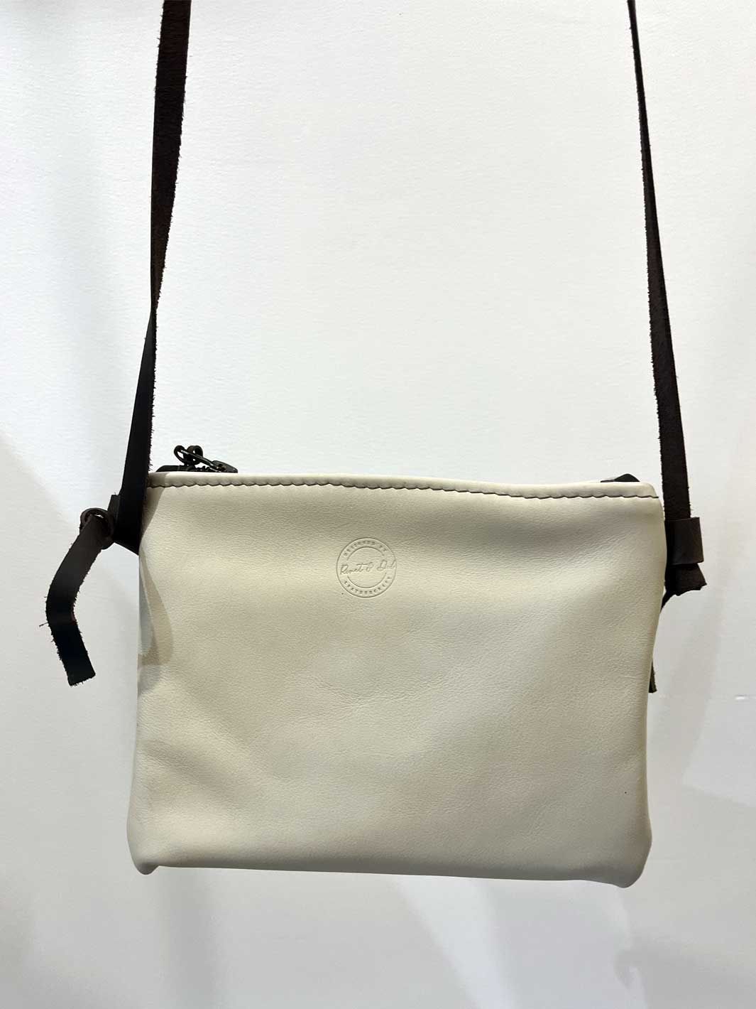 A small off-white leather crossbody bag with a dark brown shoulder strap and a debossed circular logo on the front, shown against a plain white background.