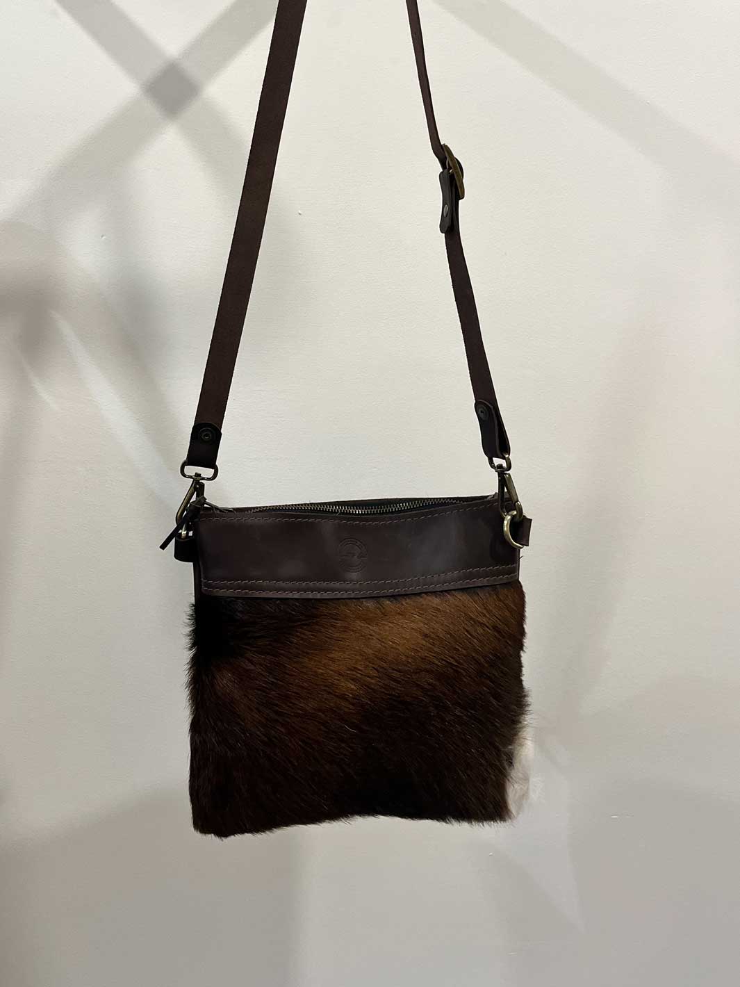 A small Australian made crossbody bag with a dark brown genuine leather top and a brown fur or hair-on-hide lower section, hanging against a white wall.
