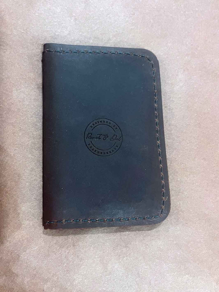 A brown leather wallet with visible green stitching and an embossed circular logo reading 'Rivet and Del' on a textured beige surface.