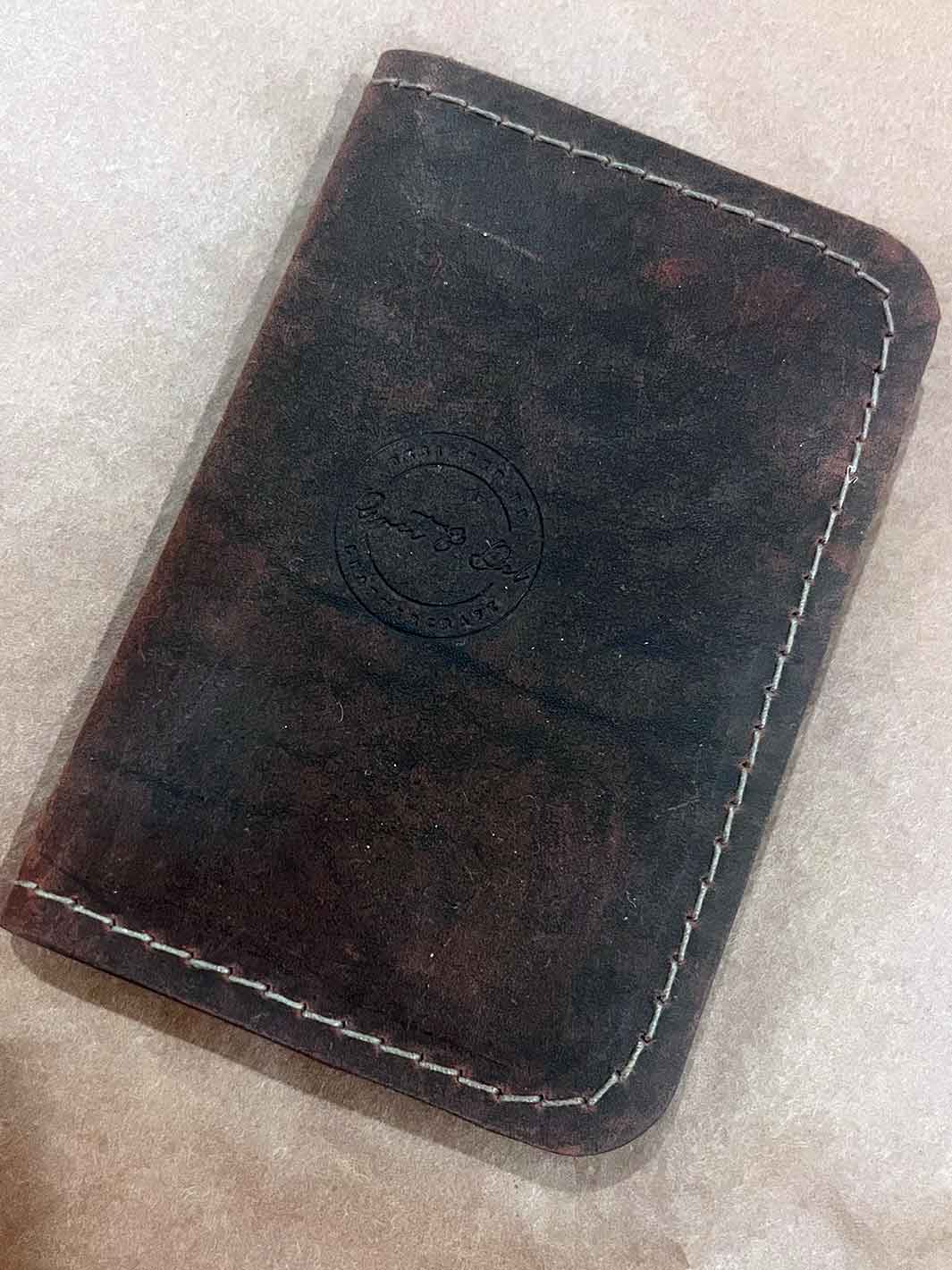 A dark brown leather card holder or wallet with brown contrast stitching and an embossed circular logo, shown on a light beige textured surface.