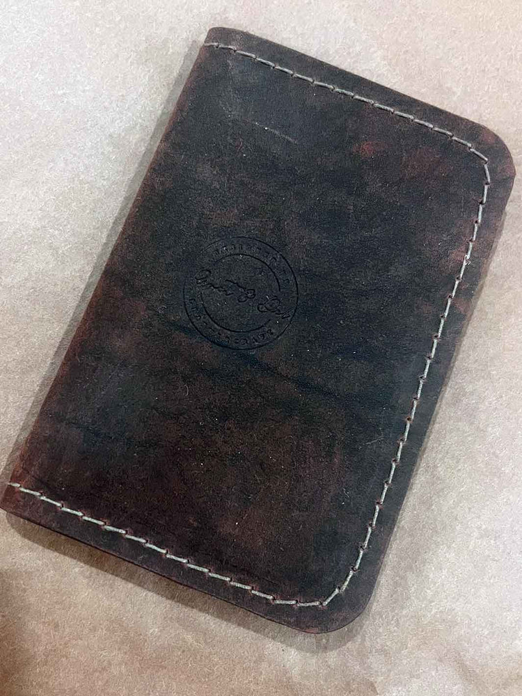 A dark brown leather card holder or wallet with brown contrast stitching and an embossed circular logo, shown on a light beige textured surface.