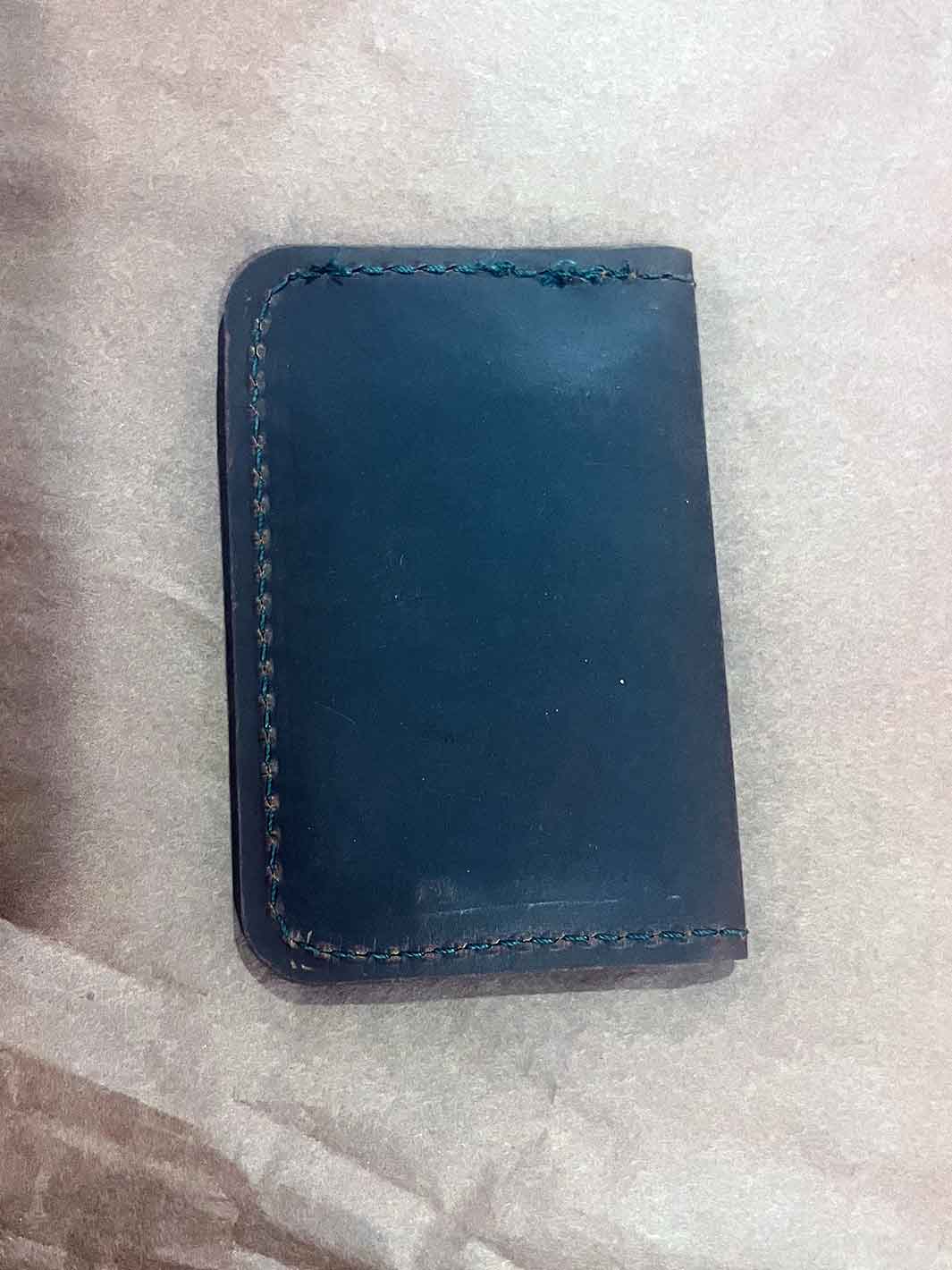 A dark brown leather card holder or wallet with visible green contrast stitching, placed on a textured beige surface.