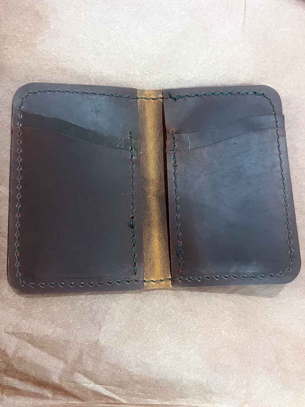A dark brown leather cardholder wallet with visible stitching and an aged effect on the fold, shown open on a light beige textured background.