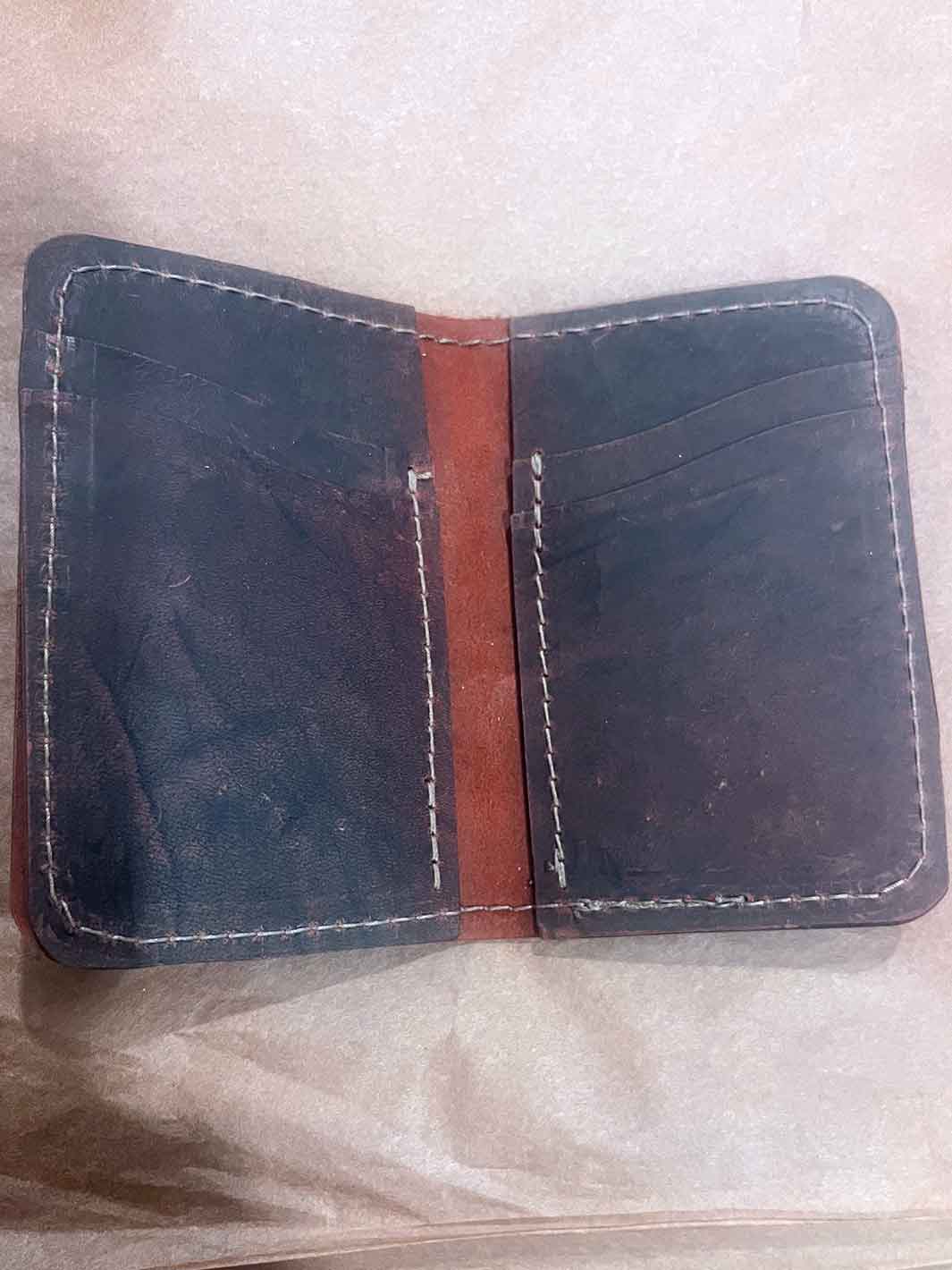 An open dark brown leather wallet with brown stitching, showing card slots and a red brown reverse side to the leather, photographed on a light beige textured surface.