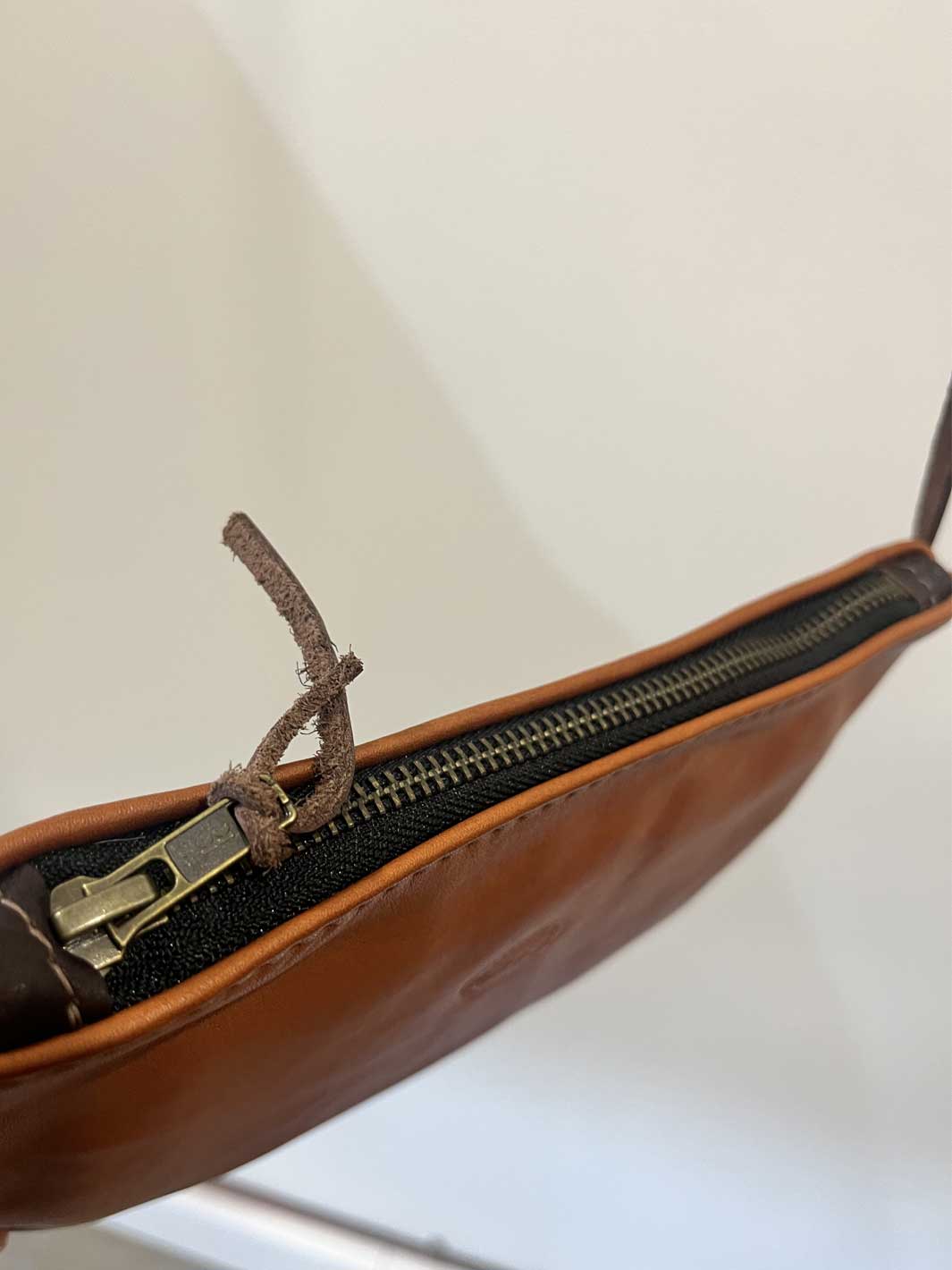 Close-up of a brown leather zippered pouch or clutch with a black zipper track and a brass-toned metal pull tab, shown against a plain light background.