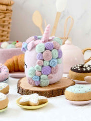 Pastel-colored unicorn-shaped cake with floral decorations on a wooden stand, surrounded by various pastries.