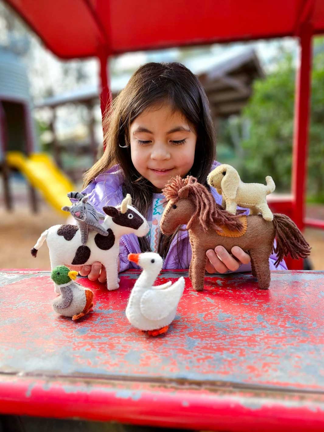 A young girl with dark hair sits at a red outdoor table, holding and playing with small felt farm animal toys including a cow, horse, sheep, duck, and goose.