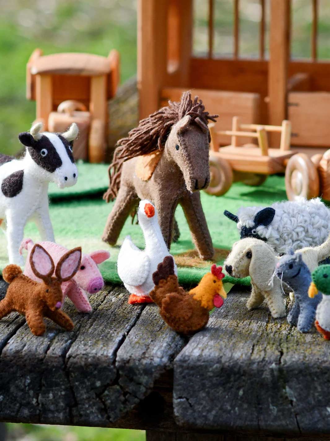 A group of small felt farm animal toys arranged on a weathered wooden surface, including a cow, horse, pig, goose, chicken, sheep, duck, and a small green bird, with a wooden toy house and cart blurred in the background.