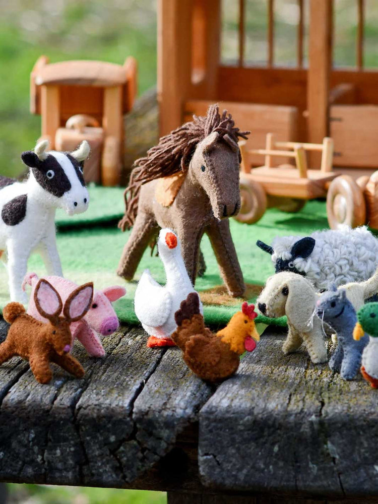 A group of small felt farm animal toys arranged on a weathered wooden surface, including a cow, horse, pig, goose, chicken, sheep, duck, and a small green bird, with a wooden toy house and cart blurred in the background.