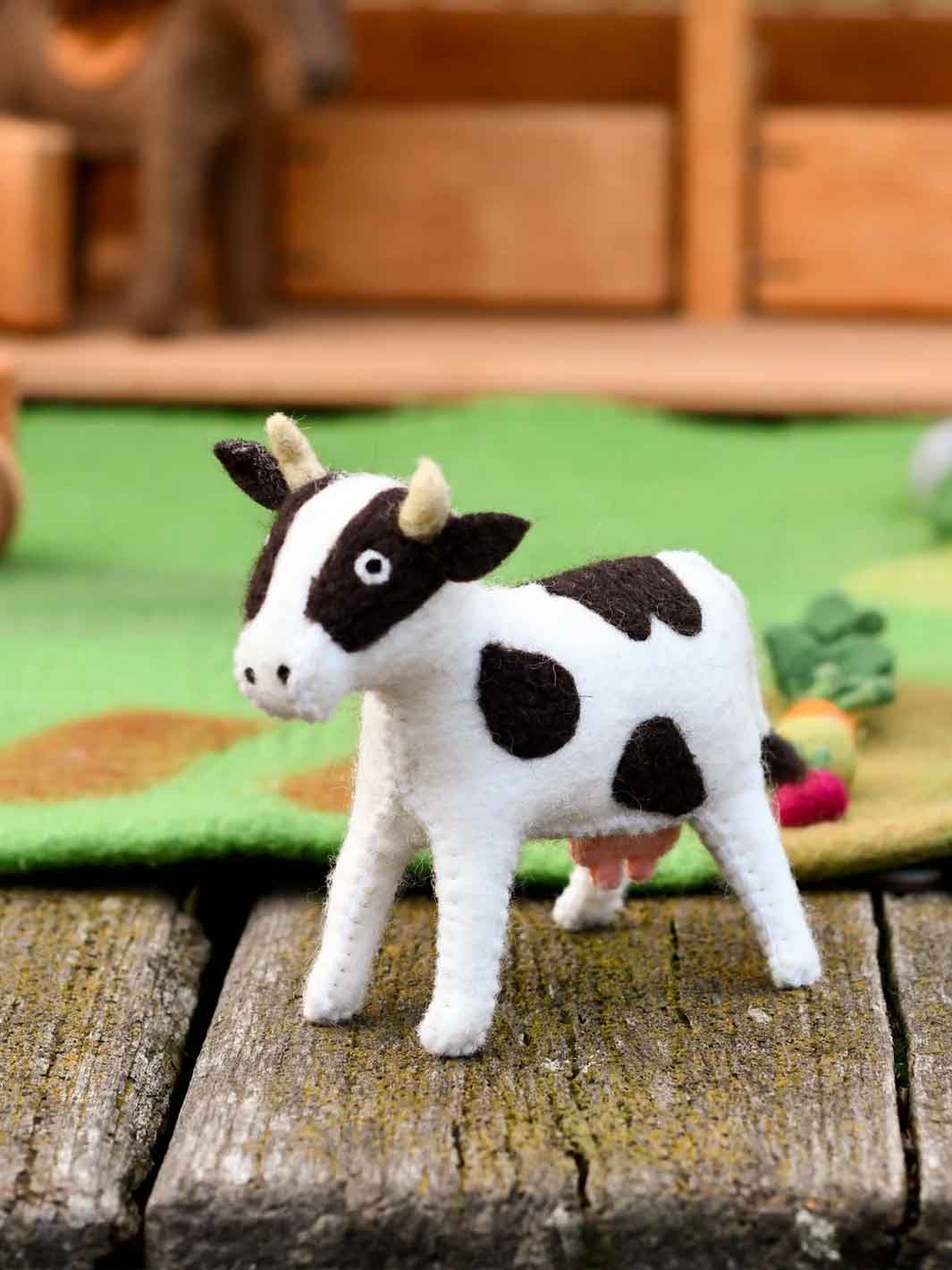 A small felt cow toy with black and white patches, standing on a rustic wooden surface with a green mat and blurred wooden background.