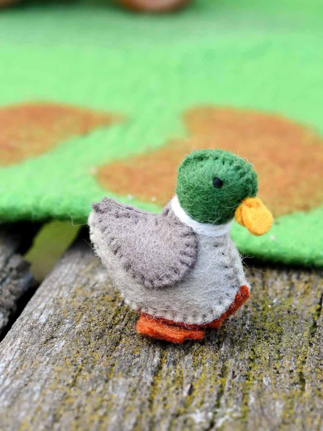 A small handmade felt duck figurine with a green head, gray body, white neck band, yellow beak, and orange feet, standing on a weathered wooden surface with a blurred green and brown background.