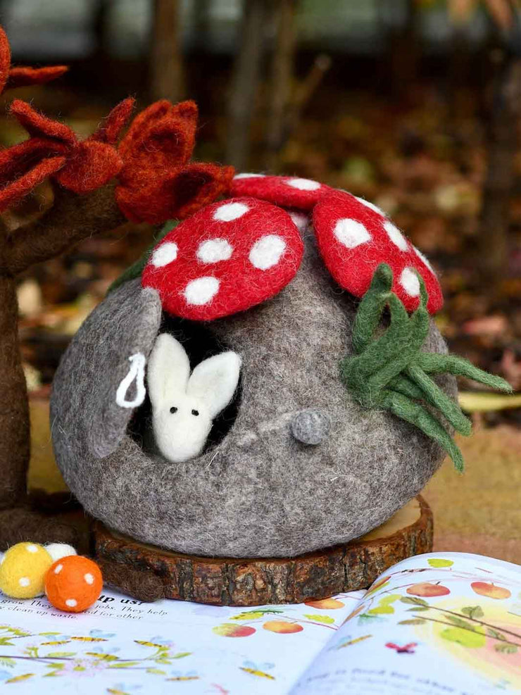 Felted mouse in a stone house with a red mushroom roof on a wooden base.