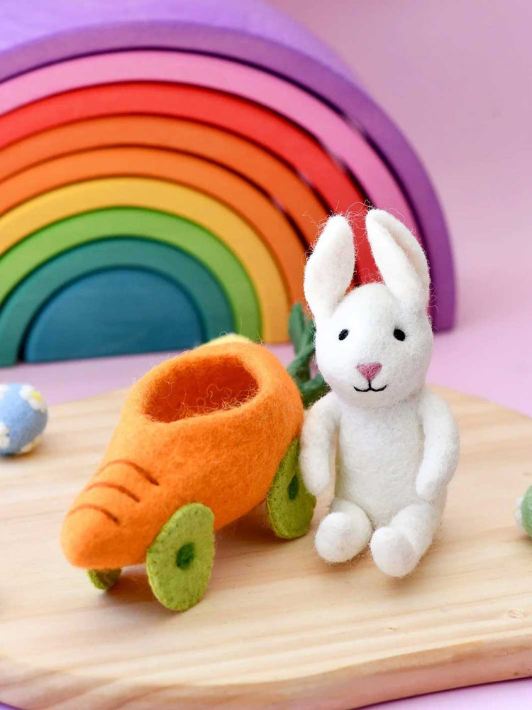 White rabbit toy with carrot-shaped car on a wooden surface with a colorful rainbow in the background.