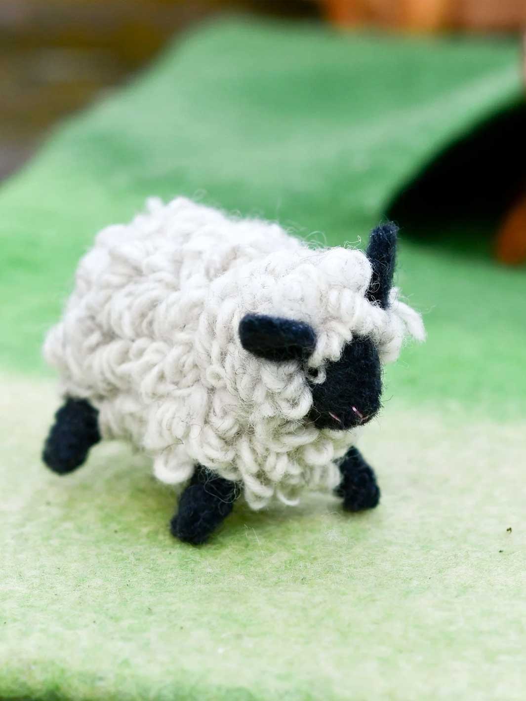 A small handmade felt sheep toy with curly white wool-like texture, a black face, and black legs, standing on a soft green felt surface.