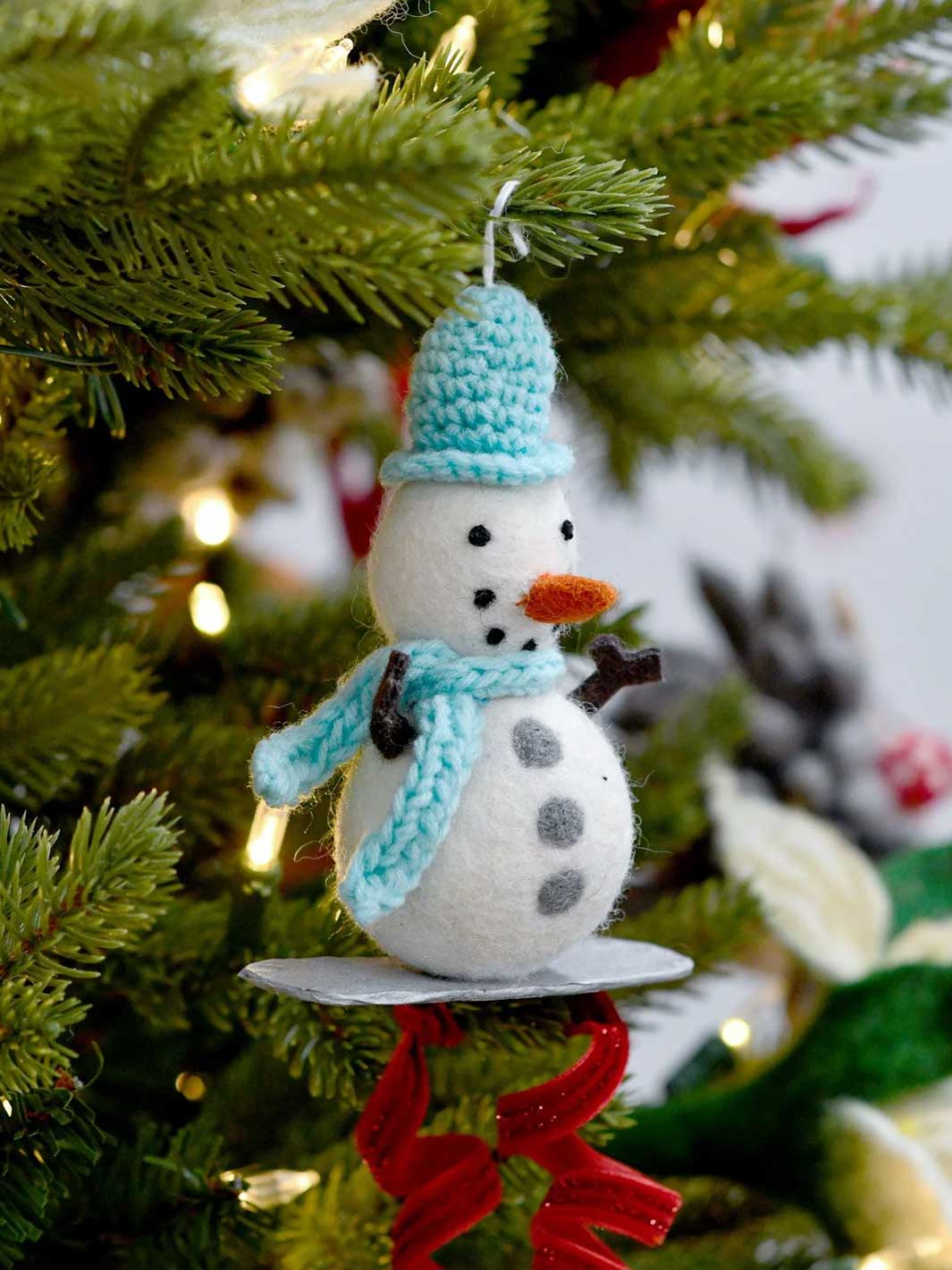 A handmade felt snowman Christmas ornament with a light blue knit hat and scarf, an orange carrot nose, and black buttons, hanging on a green Christmas tree with warm lights.