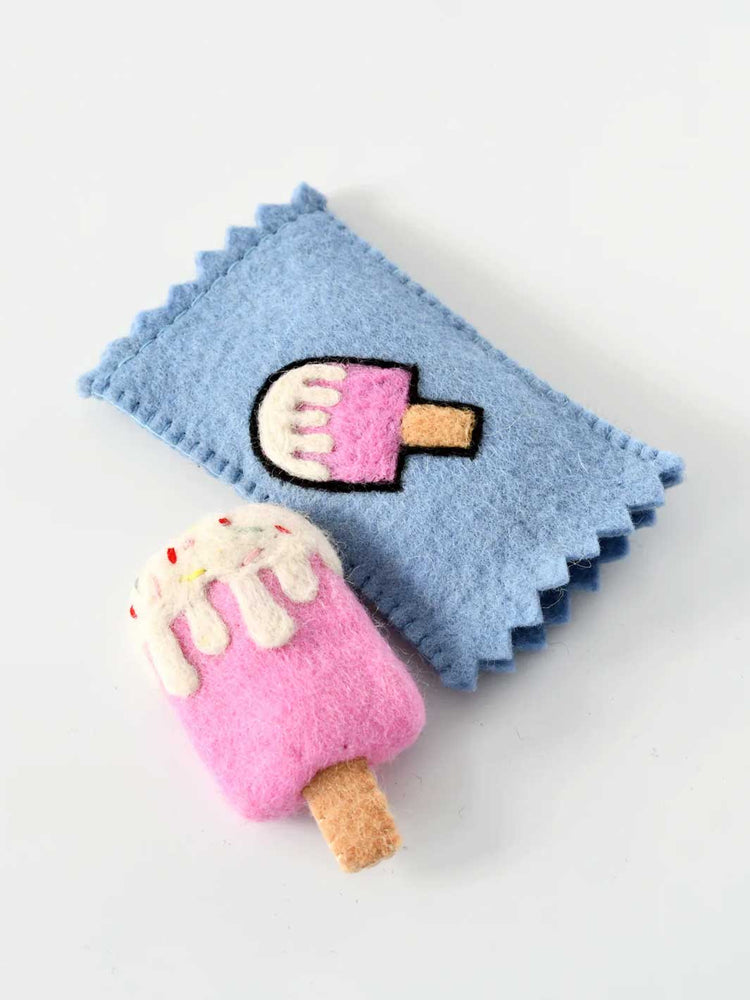 A light blue felt pouch with a scalloped edge, featuring an embroidered popsicle design, shown next to a matching pink felt popsicle charm with white frosting and a tan stick, on a plain white background.