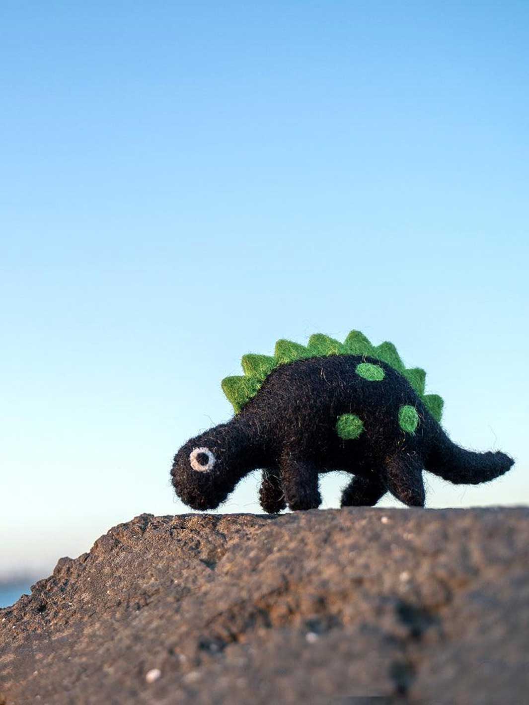 A small felted wool dinosaur toy with a black body, green spikes, and green spots, standing on a rocky surface against a clear blue sky.