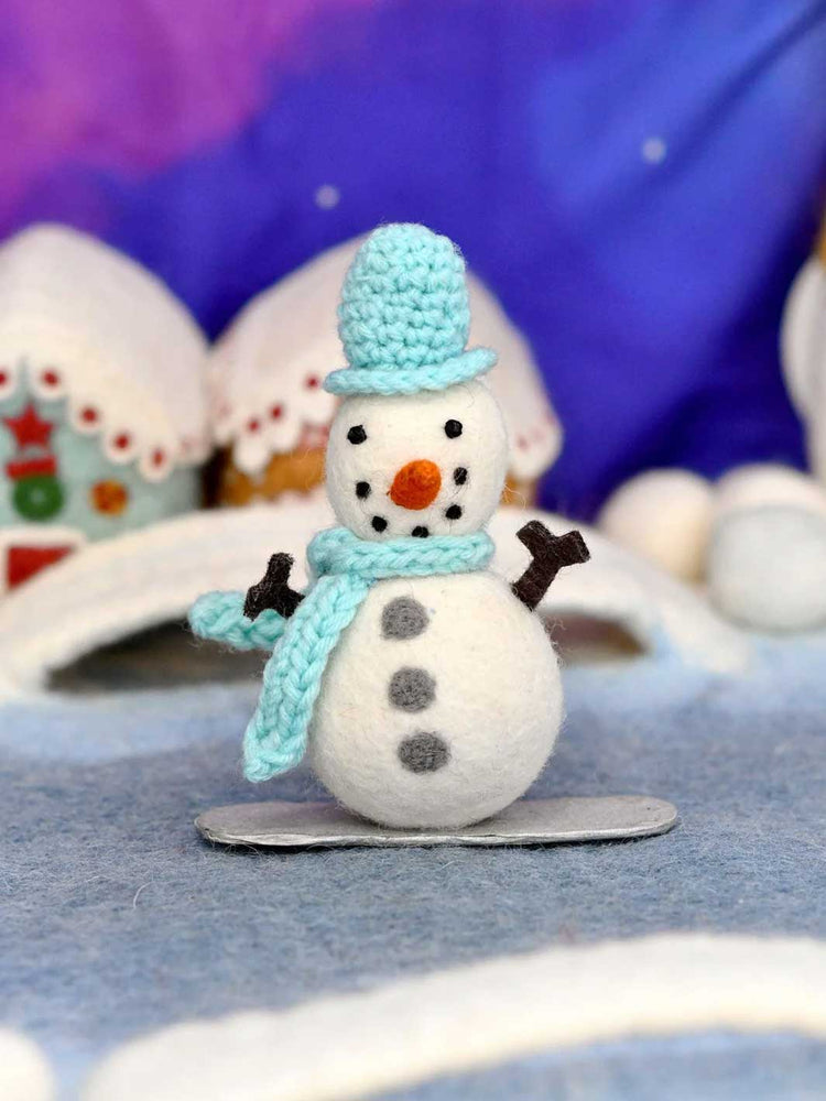 A small handmade snowman figurine made from white felt, wearing a light blue crocheted hat and scarf, with black dot eyes and arms, standing on a gray base against a blurred winter-themed background.