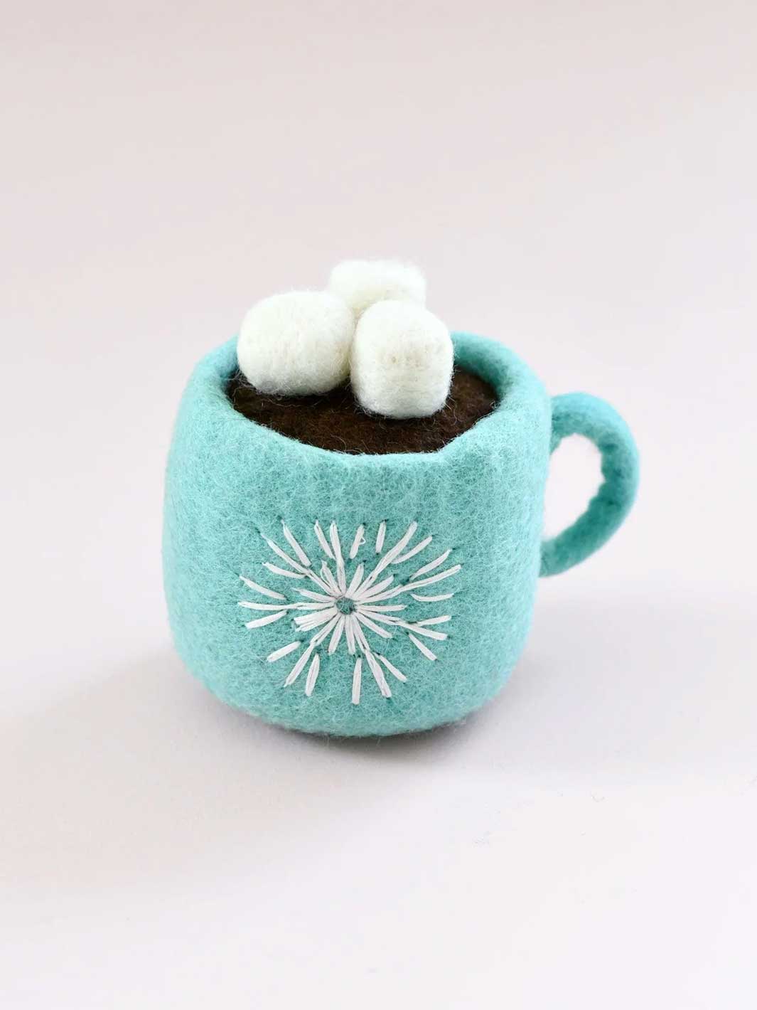 A small felt hot cocoa mug toy in light aqua blue, filled with dark brown cocoa and topped with three white marshmallows. A white embroidered snowflake design is on the front, set against a plain light background.