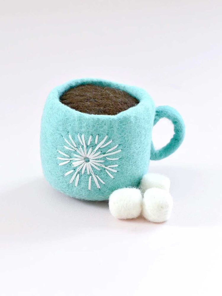 A light blue felt hot cocoa mug toy filled with dark brown cocoa, decorated with a white embroidered snowflake, with three white felt marshmallows in front, on a white background.