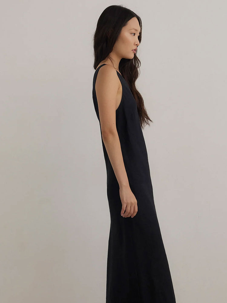 A woman wearing a sleeveless black maxi dress, shown in profile against a plain light gray studio background.