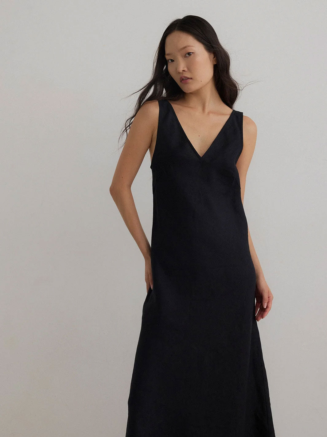 A woman modeling a sleeveless black V-neck maxi dress against a plain light gray studio background.