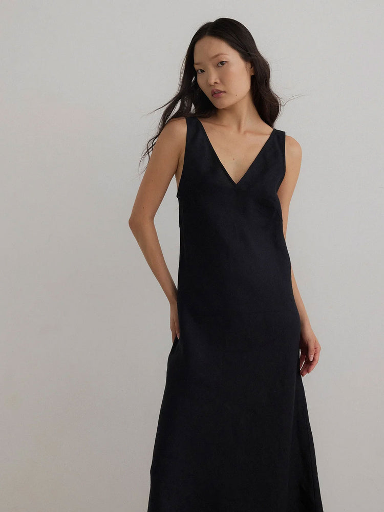 A woman modeling a sleeveless black V-neck maxi dress against a plain light gray studio background.