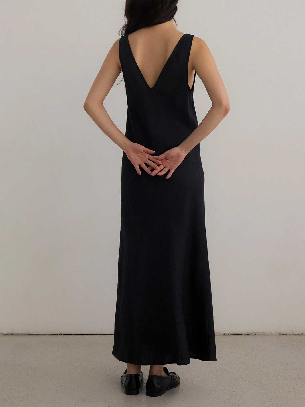 A woman viewed from behind wearing a sleeveless black maxi dress with a deep V-neckline and black flat shoes, standing against a plain light gray wall.