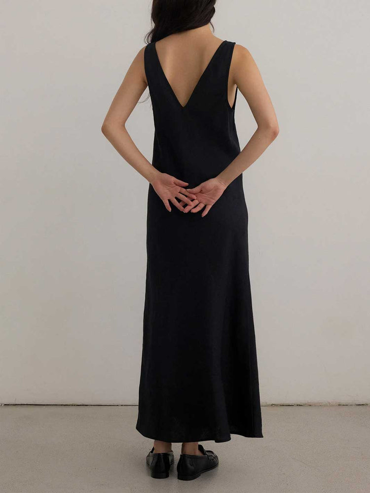 A woman viewed from behind wearing a sleeveless black maxi dress with a deep V-neckline and black flat shoes, standing against a plain light gray wall.