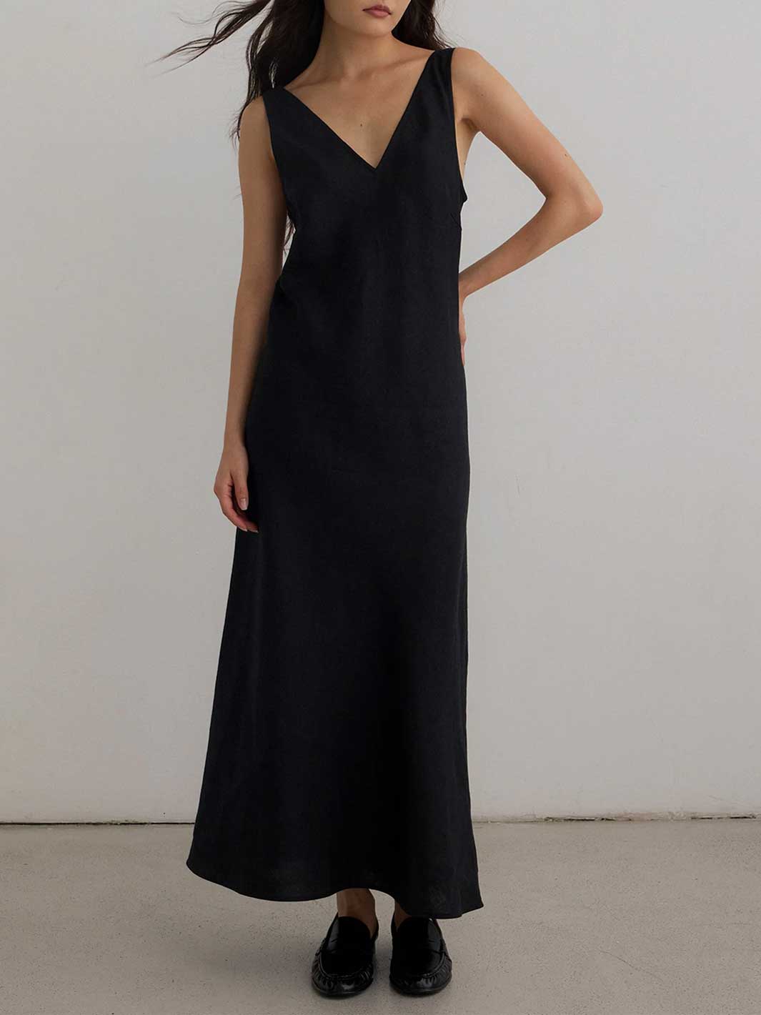 A woman wearing a long black sleeveless V-neck maxi dress with black loafers, standing against a plain light gray wall.