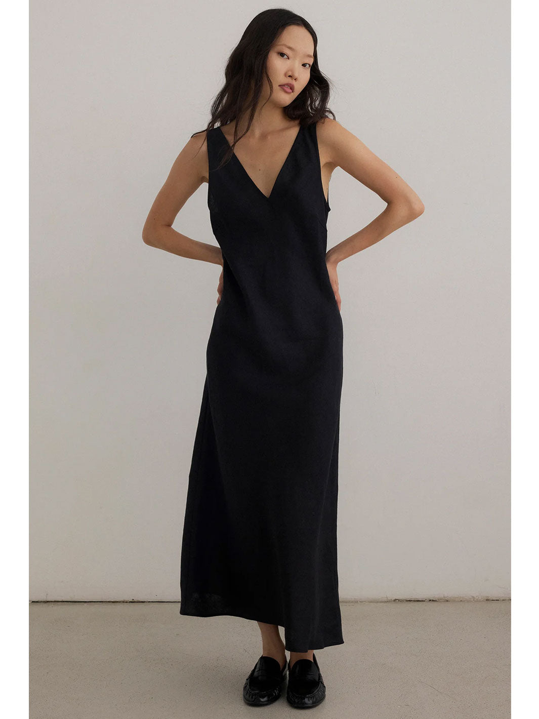 A woman modeling a sleeveless black V-neck maxi dress with a relaxed, slightly draped fit, paired with black loafers, set against a plain light gray studio background.
