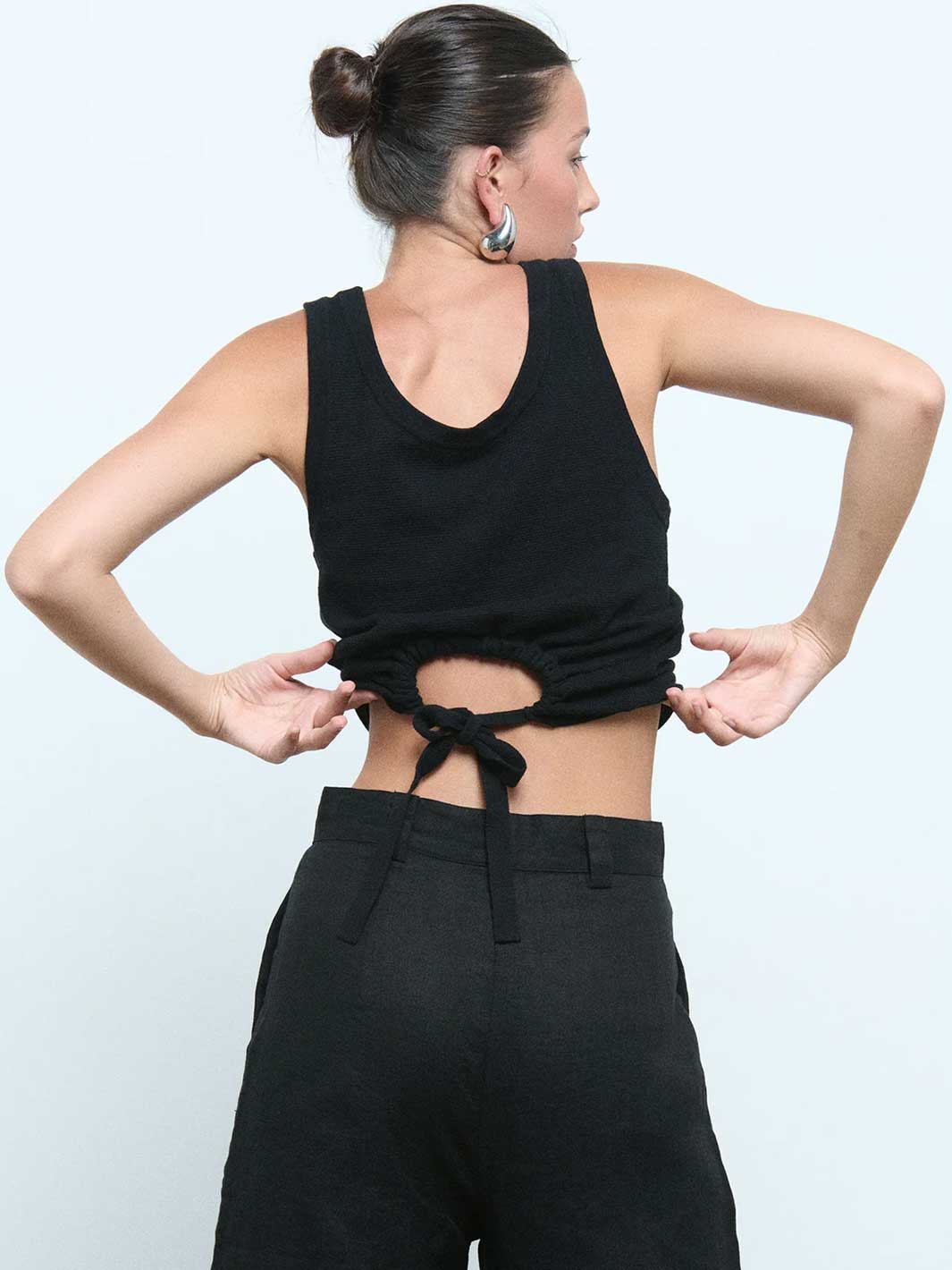 A woman shown from the back wearing a black sleeveless crop top with a cutout and tie detail at the waist, paired with black pants, against a light gray studio background.
