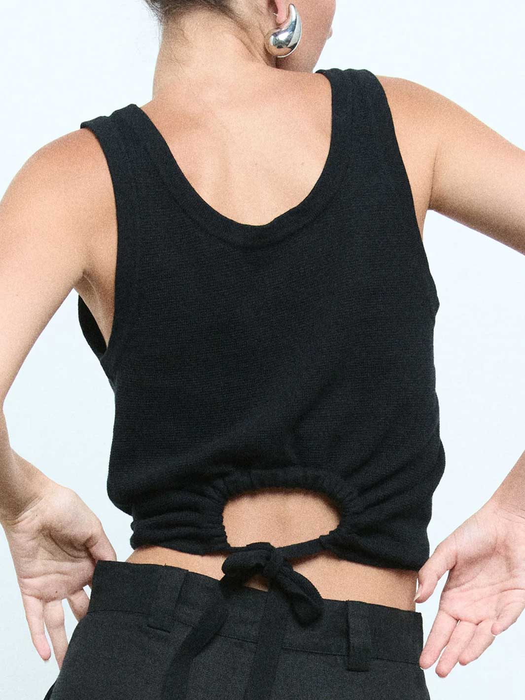A close-up back view of a person wearing a black sleeveless knit crop top with a gathered back and tie detail, paired with black pants against a light gray studio background.