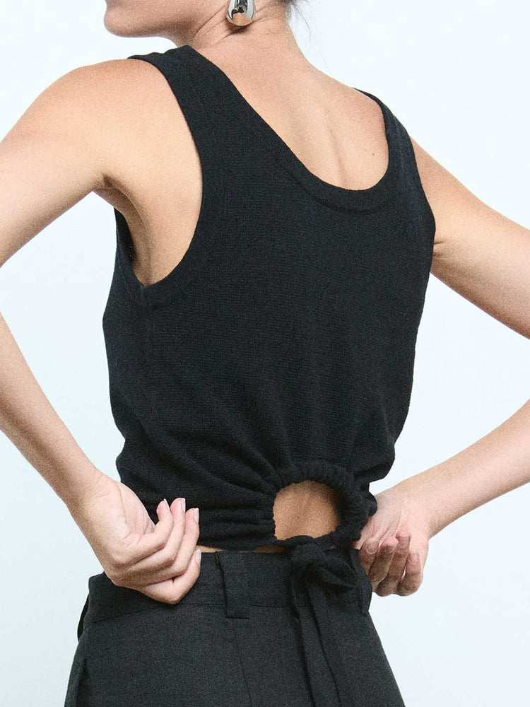 A close-up back view of a model wearing a black sleeveless knit top with a large open back and a gathered tie detail at the waist, paired with black trousers against a light gray background.